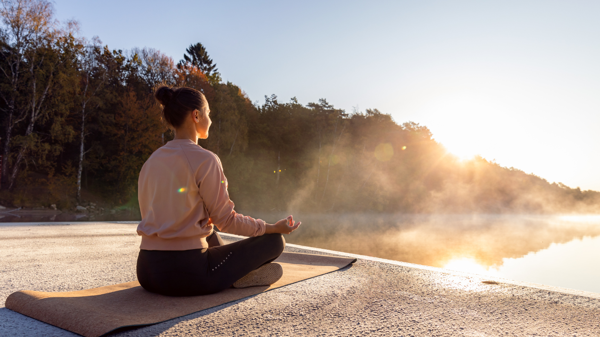 En kvinna sitter i yogaposition på en brygga med utsikt över vattnet och soluppgången.